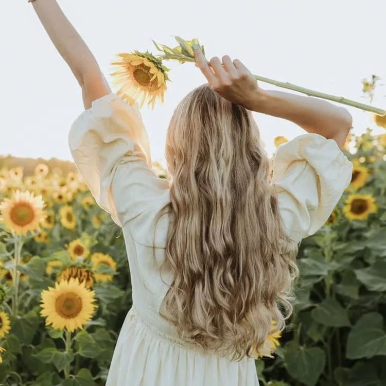 A woman with long blonde hair stands in a field of sunflowers, arms raised, holding a single sunflower.