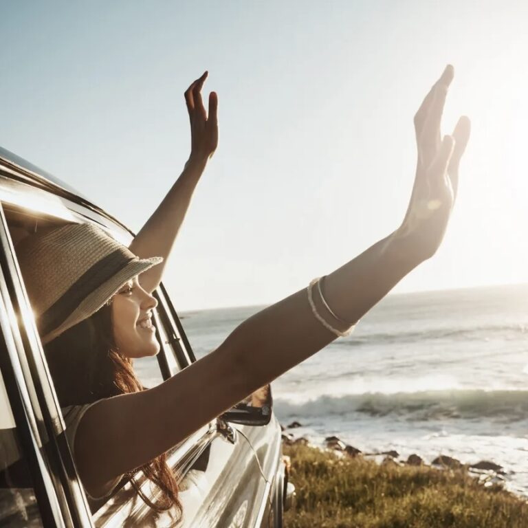 Happy woman, window and hands out with sunset in car for road trip or adventure by ocean coast in nature. Young female person enjoying journey, summer vacation or travel for drive by outdoor beach. (Happy woman, window and hands out with sunset in car