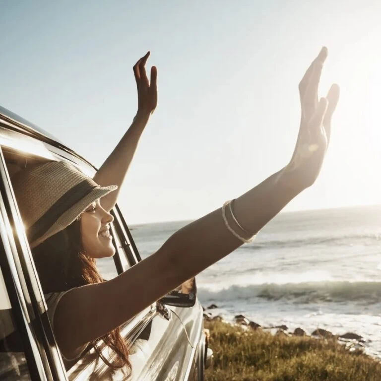 Happy woman, window and hands out with sunset in car for road trip or adventure by ocean coast in nature. Young female person enjoying journey, summer vacation or travel for drive by outdoor beach. (Happy woman, window and hands out with sunset in car