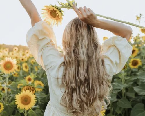 A woman with long blonde hair stands in a field of sunflowers, arms raised, holding a single sunflower.
