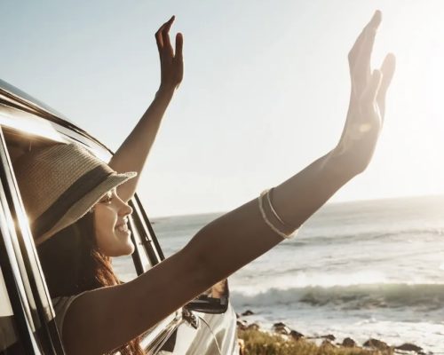 Happy woman, window and hands out with sunset in car for road trip or adventure by ocean coast in nature. Young female person enjoying journey, summer vacation or travel for drive by outdoor beach. (Happy woman, window and hands out with sunset in car